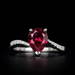 Könnte beinhalten: Ein silberfarbener Ring mit einem großen, birnenförmigen, tiefroten Edelstein in der Mitte. Die Ringschiene hat ein geschwungenes Bypass-Design und ist an beiden Seiten mit kleinen, klaren Pavé-gefassten Steinen vor schwarzem Hintergrund besetzt.