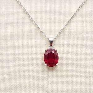 Puede incluir: Un collar de plata con un llamativo colgante de gema roja de corte ovalado. El colgante está engastado en un bisel de plata y cuelga de una fina cadena de plata. El fondo es una tela neutra de color blanquecino.