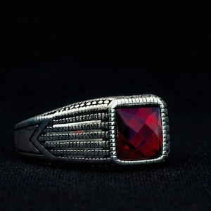 Op de afbeelding: Een zilveren ring met een grote, vierkante, gefacetteerde, dieprode edelsteen. De ring heeft een gedetailleerde, getextureerde band met geometrische patronen en een decoratieve rand rond de steen. De ring staat tegen een donkere achtergrond.