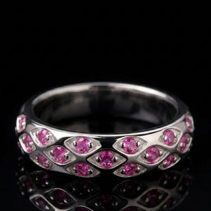 Può includere: Un anello d'argento con un motivo ripetuto di pietre preziose rosa. Le pietre sono incastonate in un design geometrico, creando un aspetto scintillante ed elegante. L'anello è esposto su uno sfondo nero.