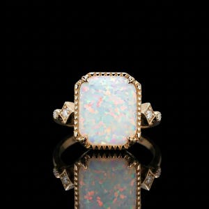 Op de afbeelding: Gouden ring met een grote, rechthoekige opaal edelsteen met een glinsterend, iriserend uiterlijk. De ring heeft een decoratieve, geschulpte rand en kleine diamantaccenten aan weerszijden, tegen een zwarte achtergrond.