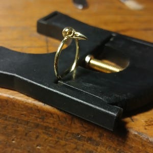 May include: A gold ring featuring a bird skull design. The ring is being held in a clamp.