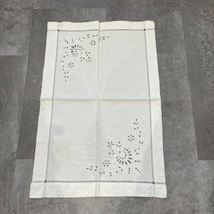 Può includere: Un runner da tavolo rettangolare in lino bianco sporco con disegni floreali e di foglie ritagliati. I bordi sono rifiniti con un bordo cucito nero. Il runner è piegato a metà, mostrando gli intricati dettagli del taglio. Misura circa 60 cm x 40 cm.