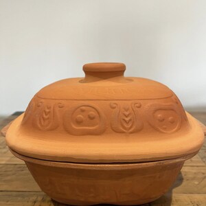 Pode incluir: Uma panela de cozinha de terracota com tampa. A panela redonda tem uma base ligeiramente alargada e uma tampa a condizer. A tampa apresenta padrões decorativos em relevo. A panela é de uma cor laranja quente e terrosa.