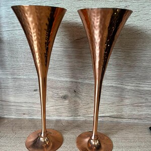 May include: Two tall, slender champagne flutes crafted from polished copper. Each flute features a distinct hammered texture on the exterior of the bowl, a long, thin stem, and a wide, circular base that provides stability for the drinkware.