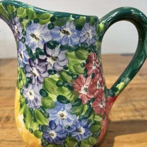 Può includere: Una brocca in ceramica dipinta a mano con un motivo floreale. La brocca ha un manico e un bordo verdi, con un corpo ricoperto di fiori blu, viola e rossi e foglie verdi. La base è gialla.