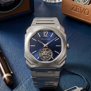 Puede incluir: Un reloj de pulsera de lujo con esfera azul y correa de metal plateado. El reloj presenta la marca "BVLGARI" y un movimiento mecánico visible. Se exhibe sobre una superficie azul, con otros relojes y accesorios en el fondo.