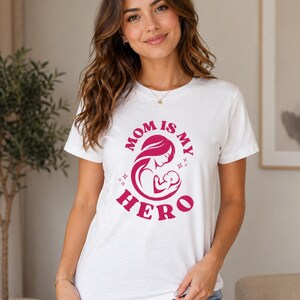May include: White t-shirt with a pink graphic that reads "MOM IS MY HERO" with an illustration of a mother and child. The shirt is a crew neck style.