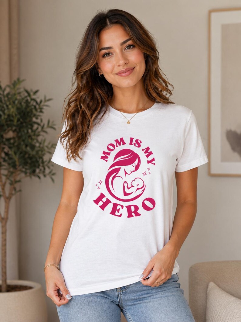 May include: White t-shirt with a pink graphic that reads "MOM IS MY HERO" with an illustration of a mother and child. The shirt is a crew neck style.