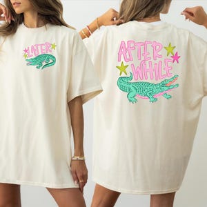 May include: Cream-coloured t-shirt with a green alligator graphic and the text "LATER" on the front. The back of the shirt features a larger alligator graphic with the text "AFTER * WHILE". The design includes pink and yellow accents.