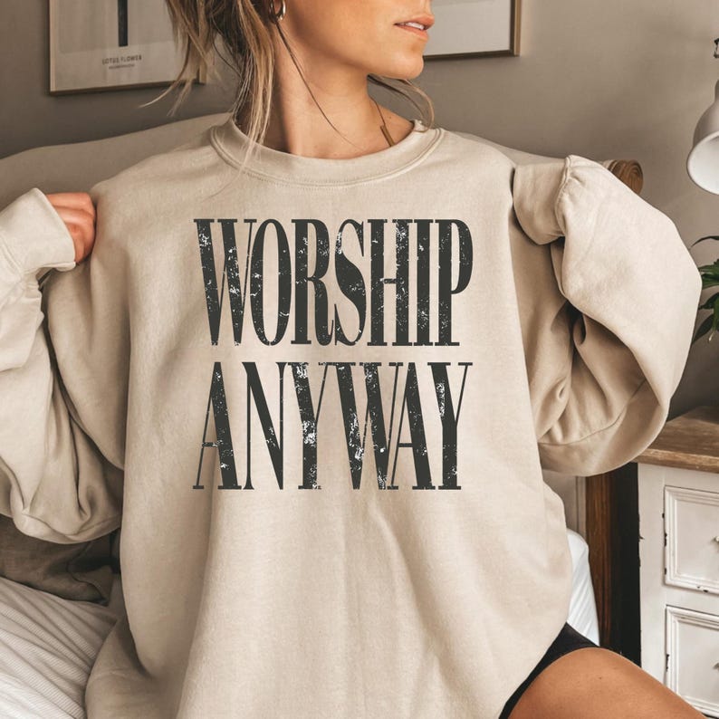 May include: A beige sweatshirt with the words "WORSHIP ANYWAY" printed in a distressed black font. The long-sleeved top is made of a soft material and is suitable for casual wear.