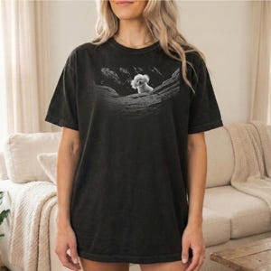 May include: Black t-shirt featuring a grayscale image of a koala bear perched on a rock formation. The koala has fluffy white fur. The shirt is a relaxed fit, with short sleeves and a crew neck.