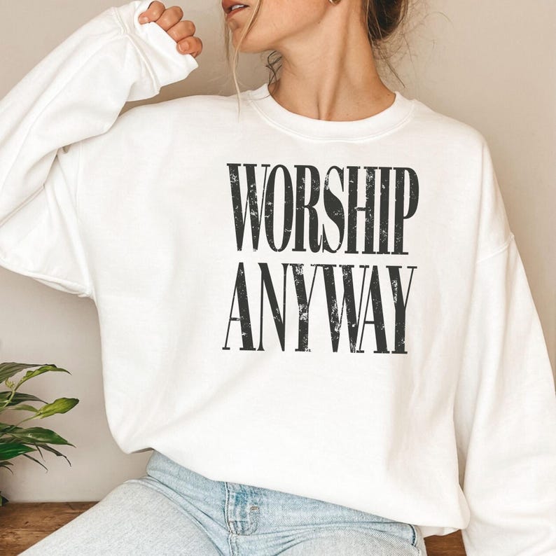 May include: White sweatshirt with the words "WORSHIP ANYWAY" printed in black, bold, capital letters. The sweatshirt has long sleeves and a crew neck. The image shows the top half of a person wearing the sweatshirt.