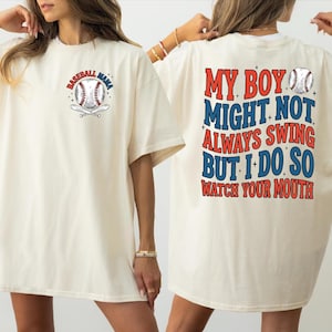 May include: Cream-colored t-shirt with baseball-themed graphics. The front features a baseball and bats with the text "Baseball Mama." The back displays the text "My Boy Might Not Always Swing But I Do So Watch Your Mouth" in red and blue.