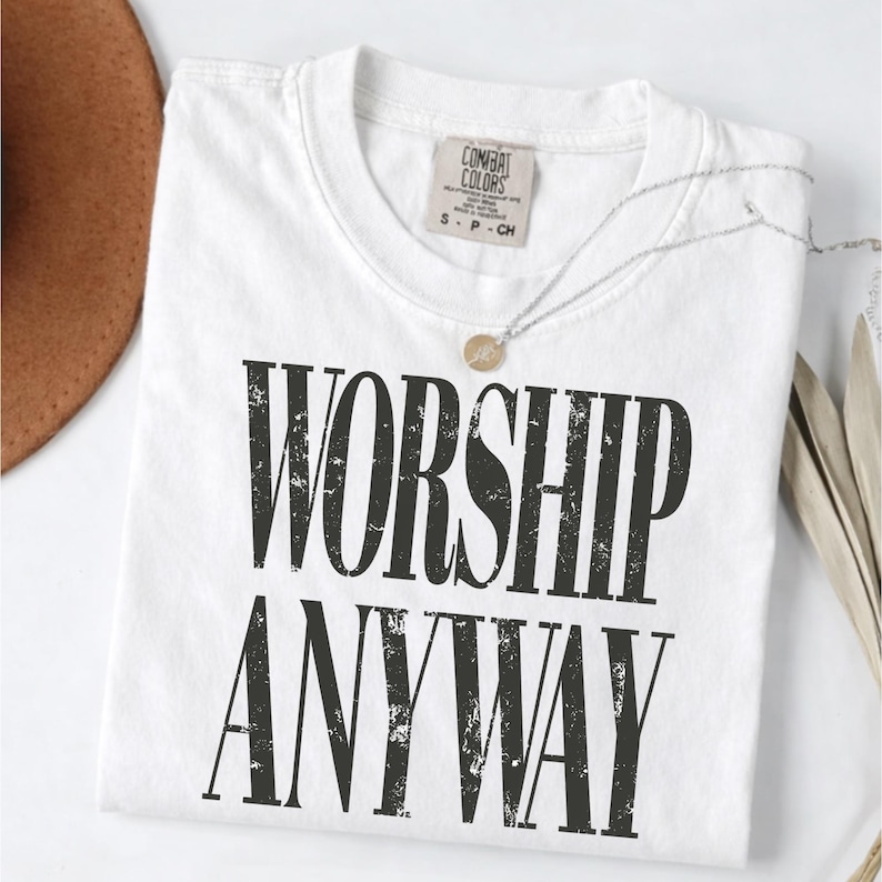 May include: A white t-shirt with the words "WORSHIP ANYWAY" printed in large, distressed black letters. The shirt has a crew neck and a small tag near the collar. A silver necklace with a small pendant is draped across the shirt.