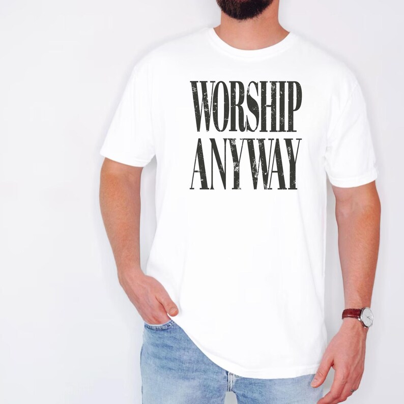 May include: White t-shirt with the text "WORSHIP ANYWAY" in a bold, black font. The shirt is short-sleeved and appears to be made of a soft material. The image shows the shirt on a person wearing blue jeans and a watch.