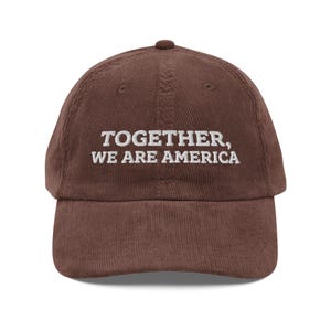 May include: A brown corduroy baseball cap with the embroidered text "TOGETHER, WE ARE AMERICA" in white. The cap has a curved brim and a small button on top. The hat is a classic style, suitable for casual wear.