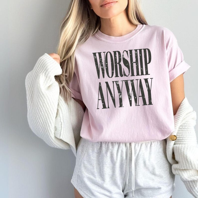 May include: A light pink t-shirt with the words "WORSHIP ANYWAY" printed in black. The shirt has short sleeves and a crew neck. The person is wearing gray shorts and a white cardigan.