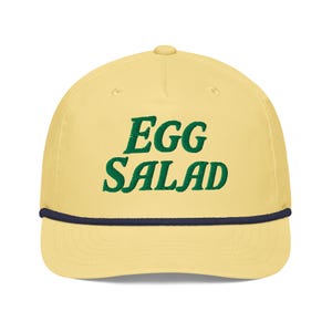 Könnte beinhalten: Hellgelbe Baseballkappe mit dem grün gestickten Schriftzug "EGG SALAD". Die Kappe hat eine marineblaue Kordel am Rand.