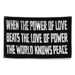 Puede incluir: Un estandarte de pared rectangular negro con el texto blanco en estilo desgastado: WHEN THE POWER OF LOVE BEATS THE LOVE OF POWER THE WORLD KNOWS PEACE. El estandarte tiene ojales metálicos en las esquinas para colgarlo, mostrando un diseño audaz.