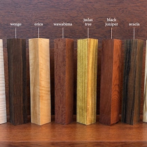 Könnte beinhalten: Acht rechteckige Holzblöcke in einer Reihe, beschriftet von links nach rechts als figured maple, wenge, erica, wawabima, judas tree, black juniper, acacia und olive. Jeder Block zeigt eine ausgeprägte natürliche Holzmaserung und Farbe.