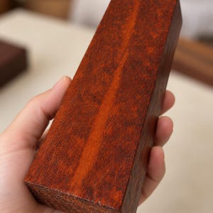 Pode incluir: Um bloco retangular de madeira castanho-avermelhada escura com um padrão de grão denso e manchado. A superfície apresenta uma cor profunda e saturada com variações subtis de tom em todo o grão da madeira, exibindo uma textura natural.