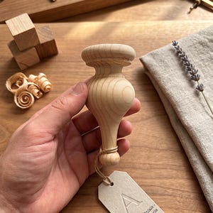May include: A turned wooden furniture foot, unfinished, held in a hand. The foot has a bulbous shape with a rounded top and a slender base. A small tag with the letter "A" hangs from the base. Wood shavings and small wooden blocks are in the background.