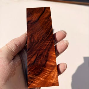 Può includere: Un blocco rettangolare di legno levigato caratterizzato da un ricco colore di base bruno-rossastro intenso con prominenti venature scure e vorticose che creano una trama complessa e fluida sull'intera superficie del pezzo di legno.