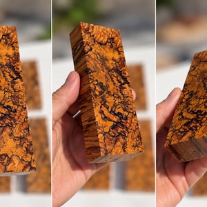 May include: A rectangular block of stabilized wood featuring a vibrant orange and golden-brown base color with intricate, dark black spiderweb-like grain patterns throughout the surface, showcasing a dense and highly figured natural wood texture.
