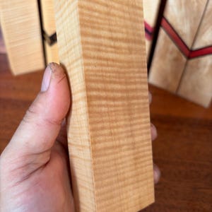 May include: A rectangular block of light-colored wood featuring a distinct wavy, tiger-stripe grain pattern throughout its surface. The wood block has a smooth, sanded finish and a natural tan hue, showcasing the organic texture of the timber grain.