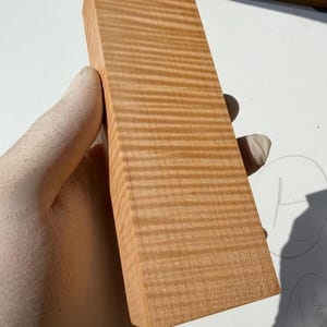 Peut inclure: Un bloc rectangulaire de bois brun clair présentant un motif de grain distinct en rayures de tigre. La surface affiche une texture naturelle, ondulée et horizontale sur toute la longueur de la pièce, mettant en valeur les détails du grain.