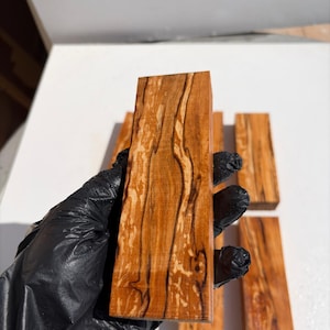 May include: A rectangular block of polished wood featuring a warm amber hue with prominent dark brown vertical grain patterns and natural swirling textures. The surface appears smooth and finished, showcasing the organic wood grain details.