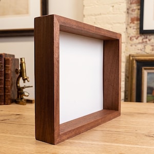 May include: A dark brown wooden picture frame with a rectangular shape. The frame has a deep profile, creating a shadow box effect. The interior is white, ready for artwork or a photograph. The frame is displayed on a wooden surface.