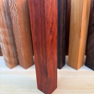 Könnte beinhalten: Eine Sammlung rechteckiger Holzkanteln für Drechselarbeiten, die aufrecht stehen. Das mittlere Stück ist tief rotbraun mit sichtbarer Maserung, flankiert von Stücken in helleren Beige-, strukturierten Brauntönen, Anthrazit und blassem Gelb.