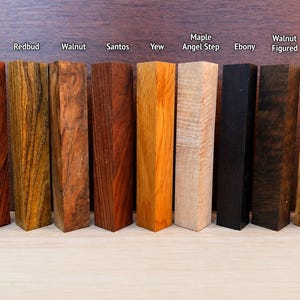 Pode incluir: Nove blocos de madeira retangulares dispostos em fila, rotulados como Bubinga, Redbud, Walnut, Santos, Yew, Maple Angel Step, Ebony, Walnut Figured e Olive Figured. Cada bloco exibe padrões de veios e cores de madeira naturais únicos.