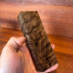 May include: A rectangular block of dark brown wood featuring a prominent, swirling grain pattern with lighter and darker streaks throughout the surface. The wood block has a smooth, polished finish and displays natural organic textures.