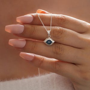 May include: A silver necklace featuring a small, almond-shaped pendant with a blue and white evil eye design. The pendant hangs from a delicate silver chain, showcasing a polished metallic finish and a centered dark blue pupil detail.