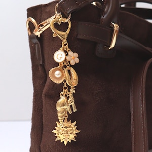 May include: A dark brown suede handbag featuring a gold-toned metal bag charm attached to the handle. The charm includes a heart-shaped clasp, a circular pendant with the letter G, a small flower, a seashell with a pearl, a giraffe, and a sun.