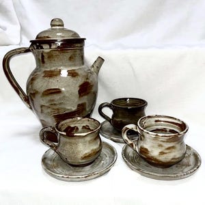Peut inclure: Service à thé en céramique fait à la main, comprenant une théière et trois tasses avec soucoupes. L'ensemble est émaillé dans des tons de gris et de brun, avec un aspect rustique et texturé. La théière a un couvercle et une anse incurvée.