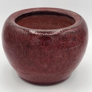 May include: A round, burgundy-colored ceramic planter with a glossy finish. The planter has a wide opening and a slightly textured surface. The color varies from a deep red to a lighter, almost purple hue in places. Ideal for indoor or outdoor use.