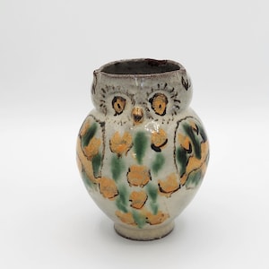 May include: A ceramic owl-shaped vase with a cream-colored base and a gray rim. The owl has yellow and green floral patterns on its body, with yellow eyes and a brown beak. The vase is a unique decorative item.