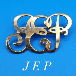 Puede incluir: Un broche de metal en tono dorado que presenta las letras en cursiva entrelazadas J, E y P. El diseño consiste en una caligrafía fluida de estilo cursivo con un acabado pulido. Las letras J, E y P son claramente visibles en el texto debajo.