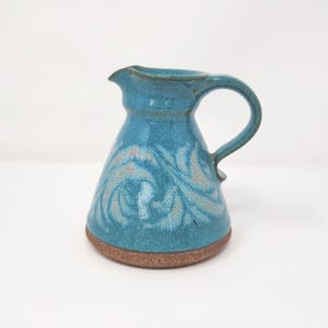 May include: A handmade turquoise ceramic pitcher with a handle and spout. The pitcher features a swirling, light-colored pattern on the body and a brown base. This decorative pitcher is suitable for serving beverages or as a decorative item.