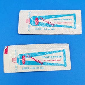 May include: Two rectangular white sample packets of Fuller Brush toothpaste. Each packet features a blue graphic of a toothpaste tube with the text Fluoride Tooth Paste with Hexachlorophene and Sample - Not for sale printed in blue and red.