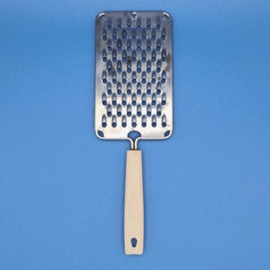 May include: A stainless steel grater with a rectangular grating surface and a cream-colored handle. The grater has numerous small holes for efficient grating. The handle is ergonomically designed for a comfortable grip. The kitchen tool is set against a solid blue background.