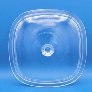 Puede incluir: Una tapa transparente de forma cuadrada con esquinas redondeadas. Tiene un borde elevado y un pequeño asa circular en el centro. La tapa es de plástico transparente y está sobre un fondo azul liso.