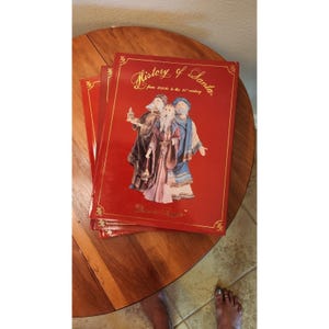 May include: A stack of red hardcover books titled "History of Santa" with gold lettering and illustrations of Santa figures. The books are on a wooden table.