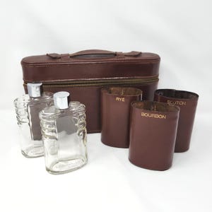 May include: A brown leather travel case with a handle and zip, two clear glass decanters with silver caps, and three brown leather-wrapped tumblers labelled with gold text reading RYE, SCOTCH, and BOURBON, arranged on a white surface.