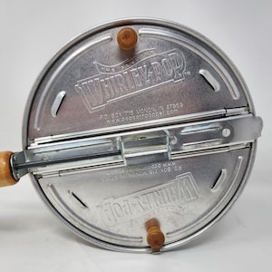 Puede incluir: Una máquina para hacer palomitas Whirley-Pop de metal plateado con mango de madera. La tapa presenta texto en relieve que dice The Genuine Whirley-Pop, P.O. Box 715 Monon, IN 47959 y www.popcornpopper.com, junto a dos perillas de madera.