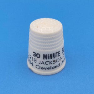May include: A white thimble with a textured surface and a slightly rounded top. The thimble has the text "30 MINUTE BE" and "2218 JACKSON St Cleveland Ave" printed on it in black. The thimble is set against a blue background.